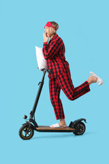 Beautiful mature woman in pajamas with pillow riding kick scooter on blue background