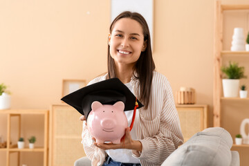 Pretty young woman holding piggy bank with graduation hat at home. Tuition fees concept - obrazy, fototapety, plakaty