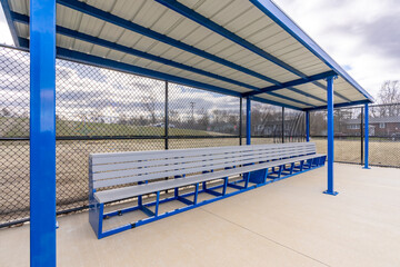 View of typical nondescript high school baseball softball dugout with concrete floor, chain link fence, and blue roof shade. No people visible. Not a ticketed event.	