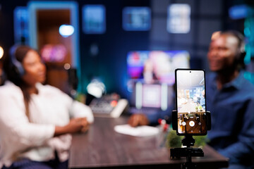 A vlogger is recording a podcast episode with a smartphone mounted on a tripod in a neon-lit...