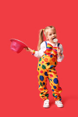 Funny little girl in clown costume with hat and lollipop on red background