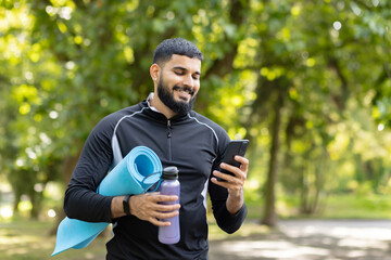 Active man with yoga mat and water bottle using smartphone in park