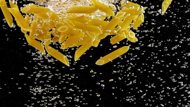 Slow Motion Close-Up Locked Down Shot: Penne Pasta Swirling Underwater with Rising Bubbles on Black Background in 4K Ultra HD Resolution
