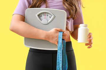 Woman in sportswear with scales, measuring tape and weight loss pills on yellow background