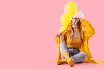 Young woman in raincoat with balloons sitting on pink background