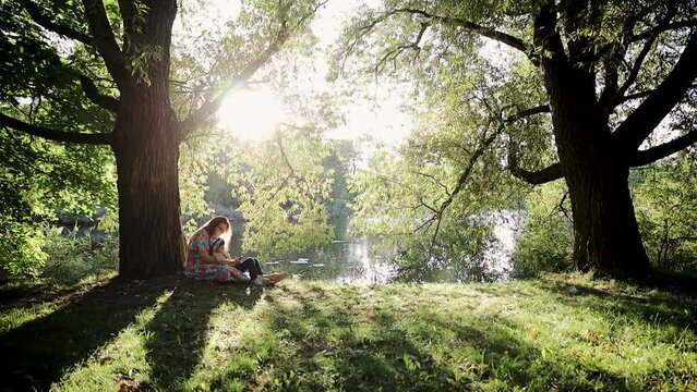 Mother with son sitting under the tree in the shadow on lawn on summer day. Steadicam shot near lake or river in park