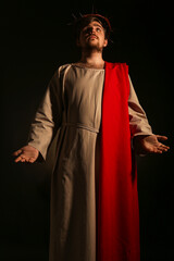 Man in Jesus robe and crown of thorns outstretching hands with light on black background