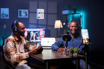 During live debate on vlog channel, happy vloggers utilizing smartphone on a tripod to film podcasts. Joyful men gathered in a studio to record broadcast using a cell phone and discuss world affairs.