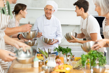 Friendly senior man, qualified chef, running culinary courses for mixed age group of ordinary passionate people, sharing professional secrets of cooking - obrazy, fototapety, plakaty