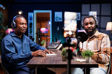 Journalist looking at camera and speaking into a microphone to introduce guest on his show. African american men with headphones are using mobile device on a tripod to record video content.
