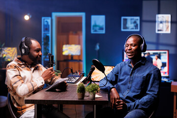 Program host and guest conversing in an elaborate home studio with neon lighting. African american influencers use professional equipment to broadcast a discussion on online podcast episode.