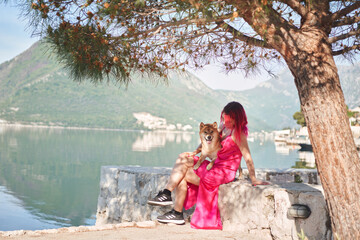 A woman with pink hair sits beside a Shiba Inu dog, both gazing out at a calm lake. The scene...