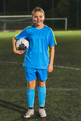 full vertical shot of a little football player girl standing with a ball in her hand, football stadium, evening. High quality photo