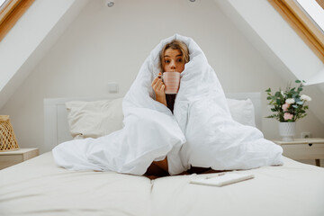 Happy woman is sitting on a bed wrapped in a white blanket and drink coffee