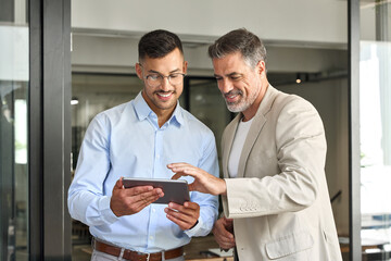 Business team of two happy successful professional business men executive working together standing in office using digital tablet computer technology managing data standing in corporate office.
