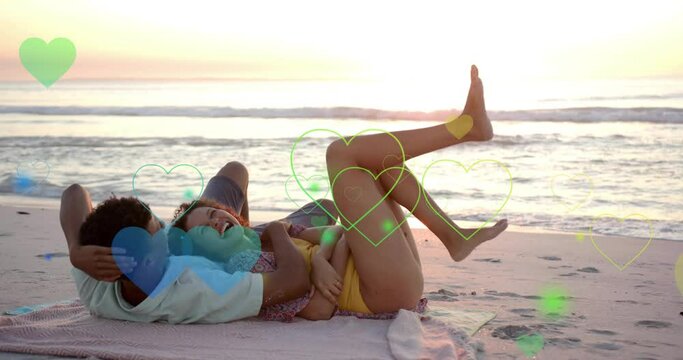 Animation of hearts moving over diverse couple in love lying on beach in summer
