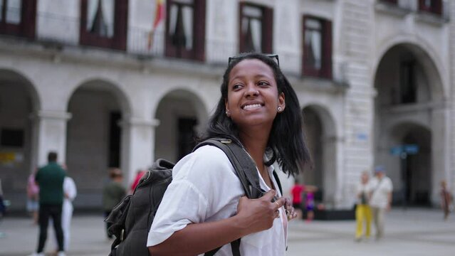 A smiling young African-American woman walks through a city square. A beautiful tourist happy as she looks around and admires the architecture of the European city