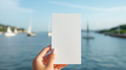 Female hand holding a piece of white paper on a blurred background of an ocean bay with yachts. Copy space for text. - obrazy, fototapety, plakaty