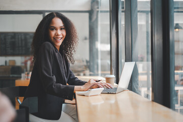 With a bright smile, a young businesswoman is engrossed in her work on a laptop at a cafe, her...