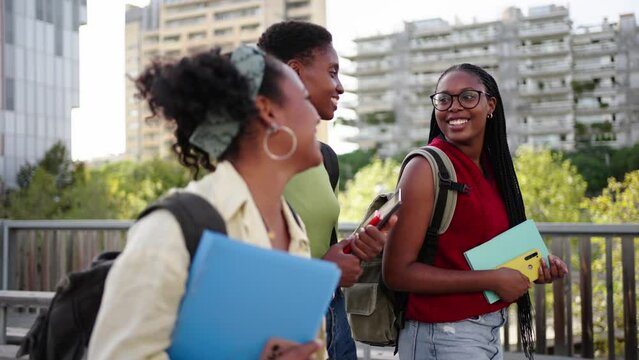Side view three African American women students strolling carefree around university campus. Young black female friends walk talking happy with backpacks and workbooks way to class sunny day outdoor