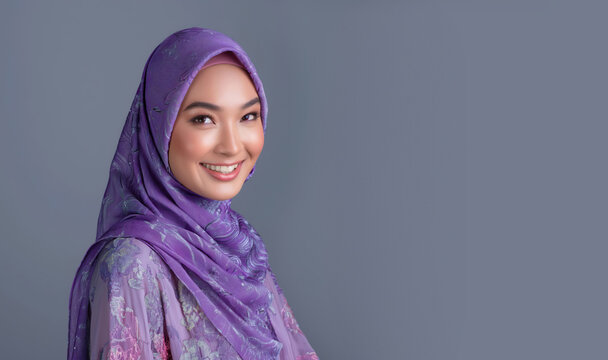 Beautiful smiling Malaysian Arab woman wearing a purple hijab and blouse with floral patterns. Grey background with copy space. An Asian girl in a traditional dress.