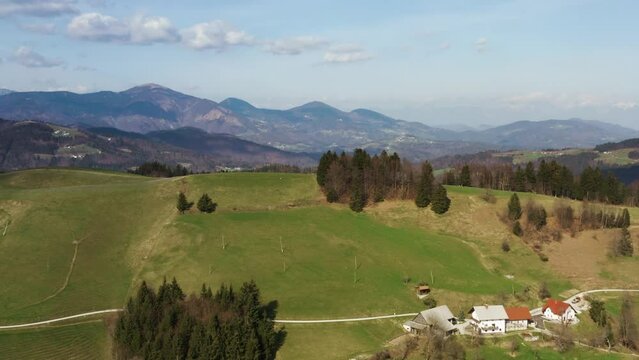 
Flying above rolling green hills with scattered trees, a rural homestead, and mountain layers fading into the horizon under a clear sky at the Mrzli Vrh.