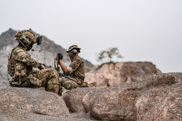 Soldiers in camo gear during a tactical operation with binoculars and rifles on a rocky terrain, showcasing military precision and readiness.