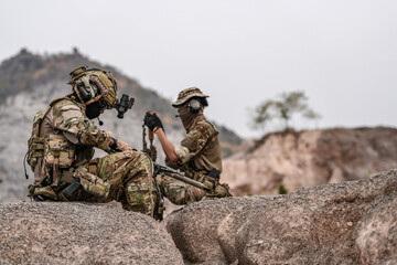 Soldiers in camo gear during a tactical operation with binoculars and rifles on a rocky terrain,...