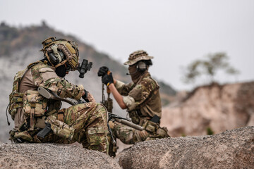 Soldiers in camo gear during a tactical operation with binoculars and rifles on a rocky terrain,...
