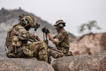 Soldiers in camo gear during a tactical operation with binoculars and rifles on a rocky terrain,...