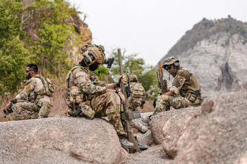 Soldiers in camo gear during a tactical operation with binoculars and rifles on a rocky terrain,...