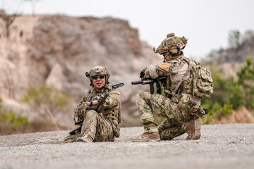 Soldiers in camo gear during a tactical operation with binoculars and rifles on a rocky terrain,...