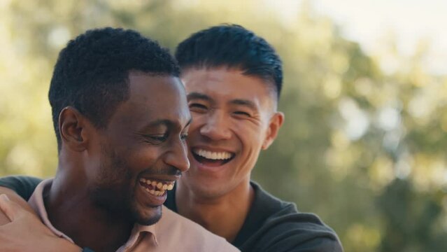 Close up portrait of same sex male couple or friends standing outside in beautiful countryside laughing and smiling as they hug and look at camera - shot in slow motion