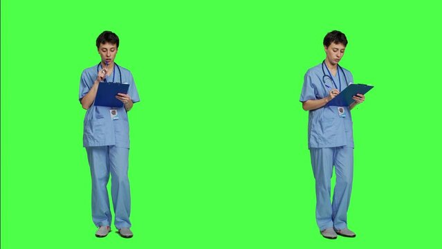 Medical assistant writing checkup information on clipboard files, taking notes and making doctor appointments with checklist. Nurse wearing blue scrubs stands against greenscreen backdrop. Camera A.