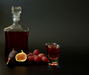 Fig-grape liqueur in a bottle and two glasses on a black background, next to ripe fruits.