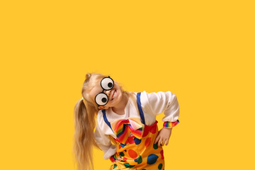 Playful little girl in clown costume with funny glasses on yellow background