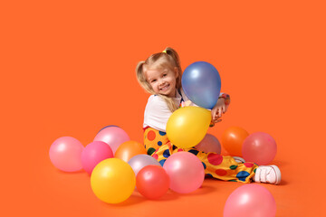 Fototapeta na wymiar Smiling little girl in clown costume with balloons on orange background