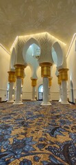 The interior of the grand mosque  has white and gold columns with a blue carpet featuring a floral...