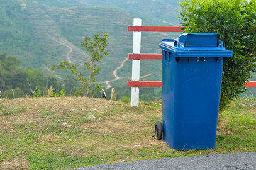 Beside the hill, on the roadside, sits a plastic blue garbage container.