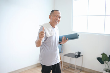 Fototapeta na wymiar Senior Asian man holding rolled mat and showing thumbs up after exercise at home