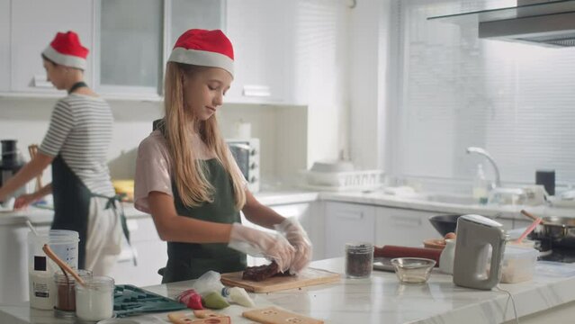 Pan shot of teen girl in Christmas hat making chocolate dough for cookies when helping mother at home kitchen