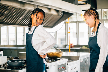 Girlfriend chef helps woman friend tie apron in a restaurant kitchen. Two adults in uniform prepare for food education and service in the industry.