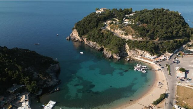 Aerial estabilshing orbit above golden sandy beach and cove in Corfu Greece with boats at dock and tourists on beach