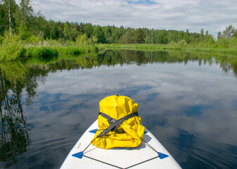 summer landscape from the river, in the foreground a sup board and a yellow rubber bag, cloud reflections in the water, green trees and grass on the banks of the river, Sedas River, Latvia