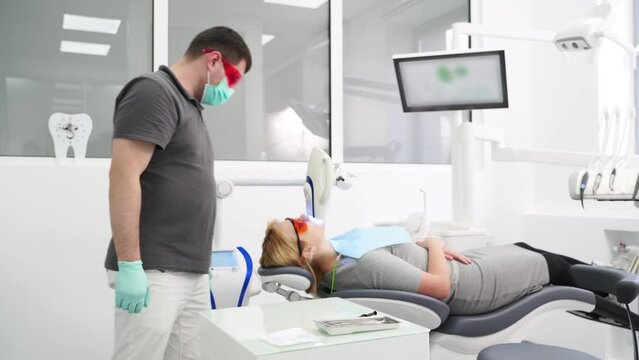 Professional UV whitening teeth procedure in the modern dental clinic. Male dentist checking female patient during laser teeth bleaching. Blonde woman in protective red glasses. Cosmetic dentistry.