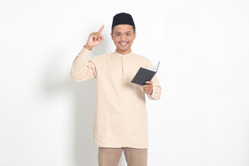 Portrait of attractive Asian muslim man in koko shirt with peci reading a book, telling that he has an idea while pointing finger and pen. Isolated image on white background