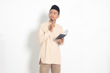 Portrait of confused Asian muslim man in koko shirt with peci reading a book, thinking about an idea with hand holding pen on chin, looking away. Isolated image on white background