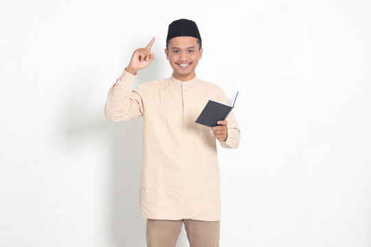 Portrait of attractive Asian muslim man in koko shirt with peci reading a book, telling that he has an idea while pointing finger and pen. Isolated image on white background