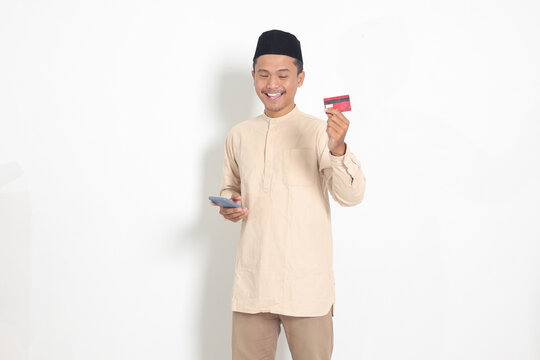 Portrait of attractive Asian muslim man in koko shirt with skullcap holding a mobile phone and presenting credit card. Isolated image on white background