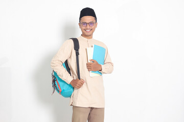 Portrait of excited student Asian muslim man in koko shirt with skullcap carrying backpack, while...